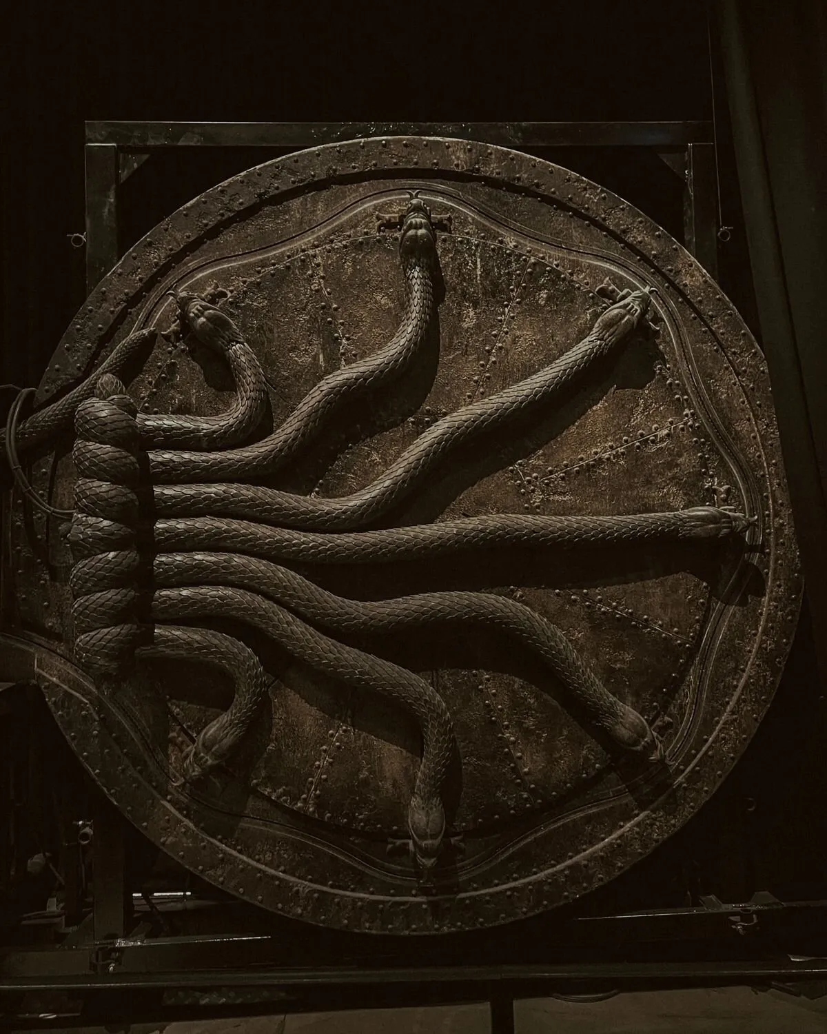 A porta redonda da Câmara Secreta, de metal escuro e cheia de rebites, com várias serpentes esculpidas que saem de um feixe enrolado à esquerda e se esticam até a borda, cada uma com a cabeça presa a um ferrolho
